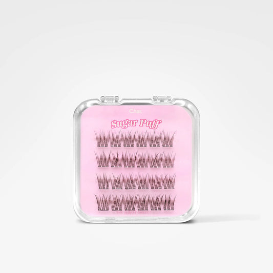 A clear plastic clamshell case containing false eyelash clusters on a pink backing card labeled 'Sugar Puff' and 'Clás', photographed on a clean white background.