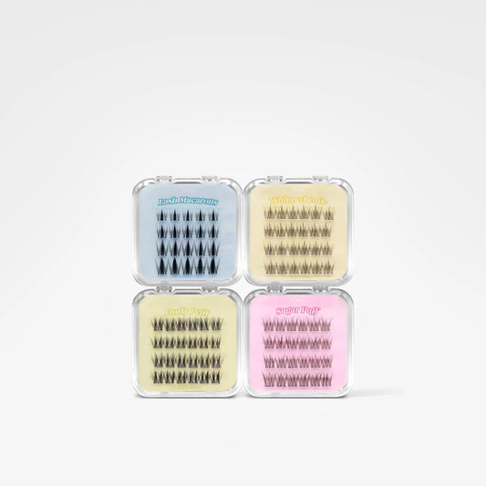 Four small clear plastic cases containing false eyelashes, arranged in a 2x2 grid on a light gray background. The cases are colored light blue, pale yellow, cream, and pink, with the brand name 'Peachy Pig' visible on the top of each case.