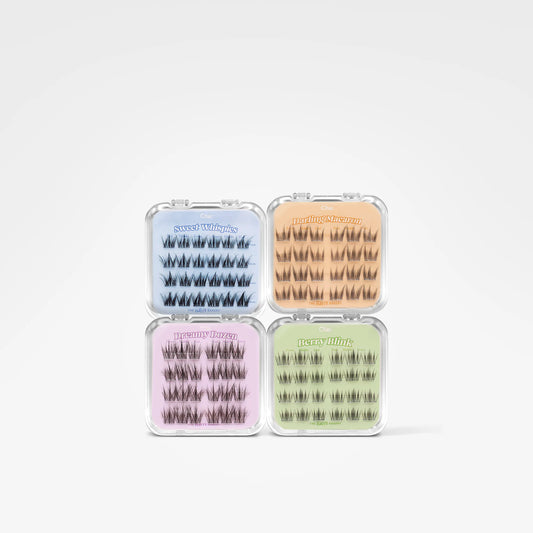 Four small clear plastic cases containing false eyelashes, arranged in a 2x2 grid on a light gray background. The cases are colored light blue, peach, pink, and sage green, with the brand name 'Barely Visible' printed on each.