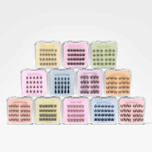Twelve small clear plastic cases containing false eyelashes, arranged in a 3-by-4 grid on a light gray background. The cases come in assorted pastel and neutral shades including pink, yellow, blue, green, and beige.